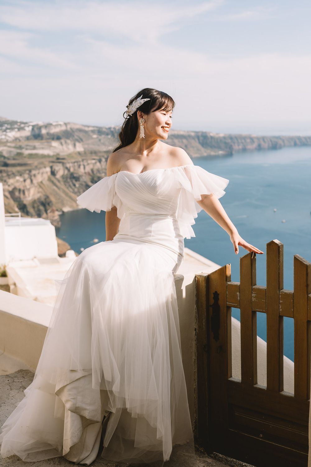 Smiling bride in white off-shoulder gown poses by wooden gate with Santorini cliffs and blue sea backdrop.