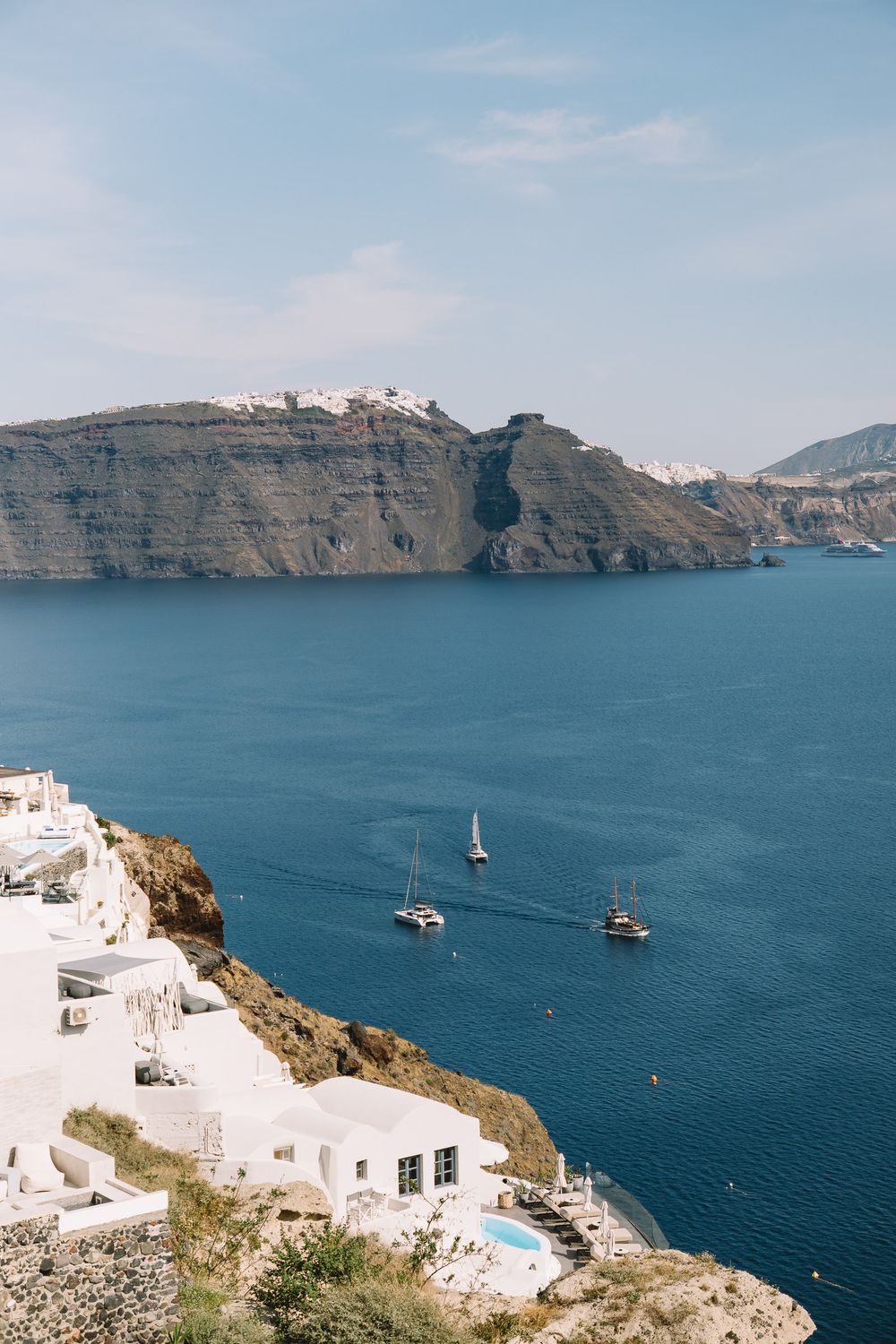 Stunning aerial view of Santorini caldera with white-washed buildings on cliffs, sailboats on blue Aegean Sea, and volcanic cliffs.