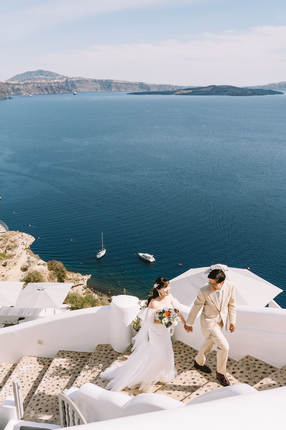 Bride and groom walk hand-in-hand on white steps in Santorini, Greece, with stunning caldera sea views.