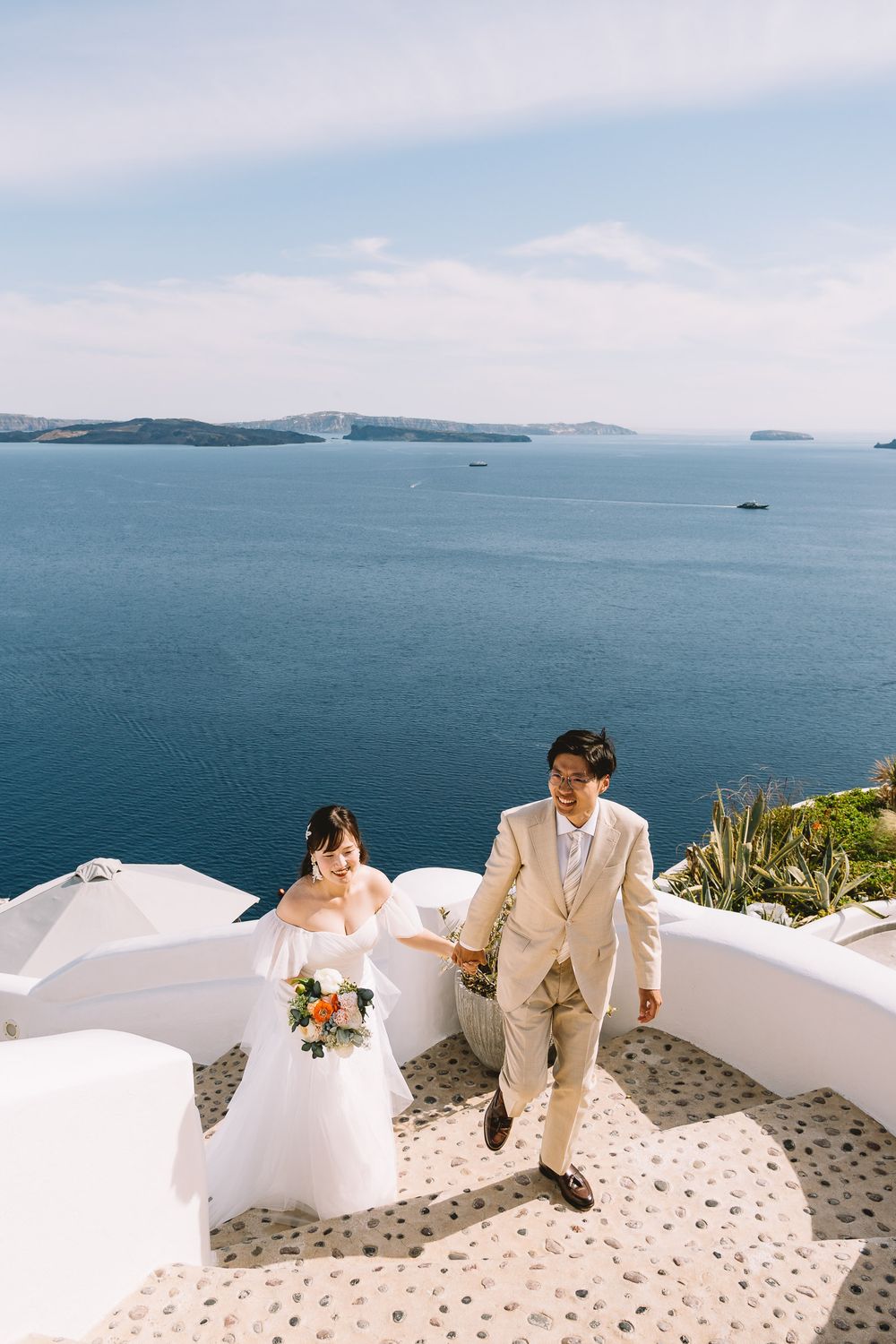 Couple in wedding attire holding hands on white steps in Santorini, Greece, with stunning blue Aegean Sea backdrop.