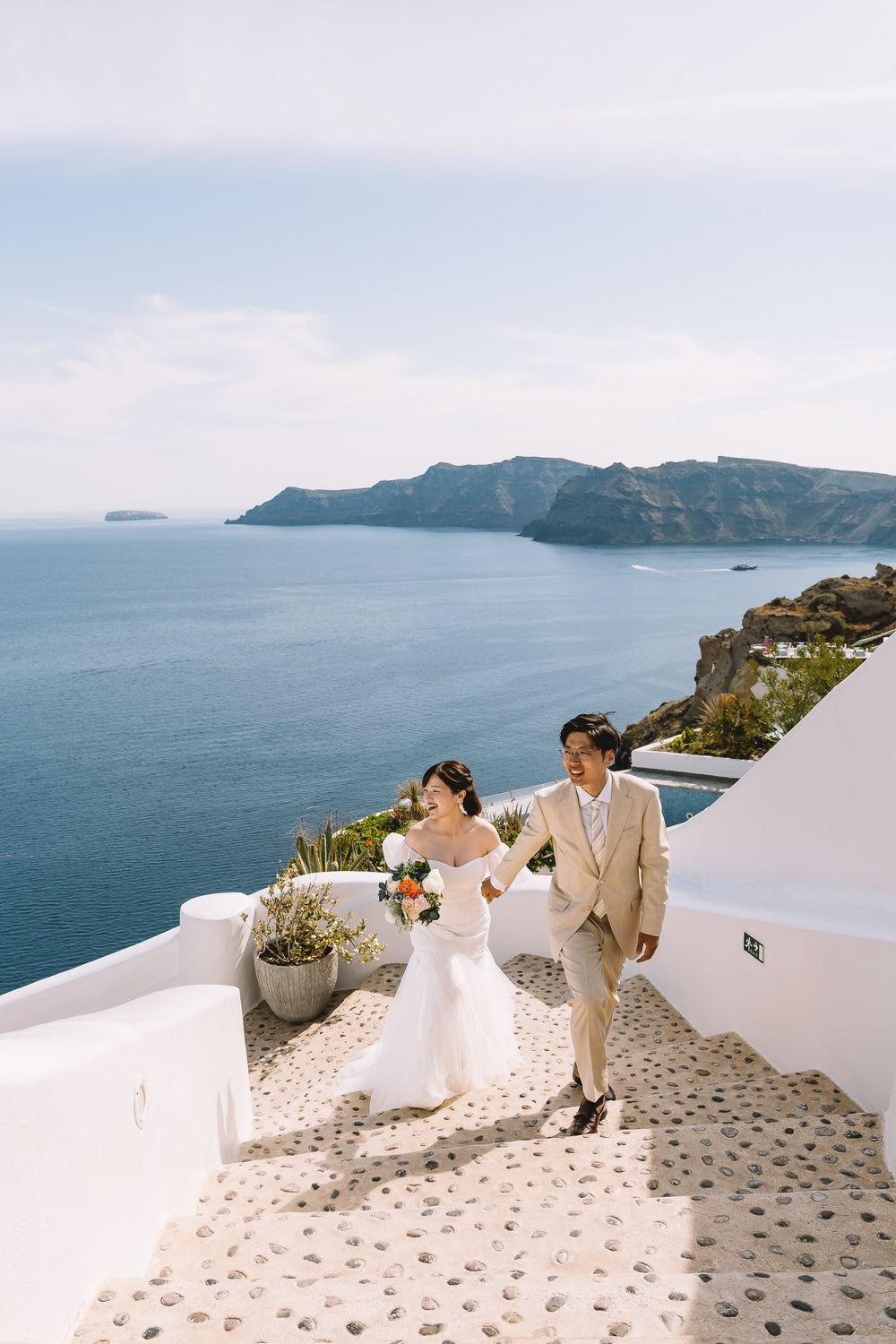 Couple in wedding attire walks along white Santorini steps with stunning blue Aegean Sea and cliffs backdrop.