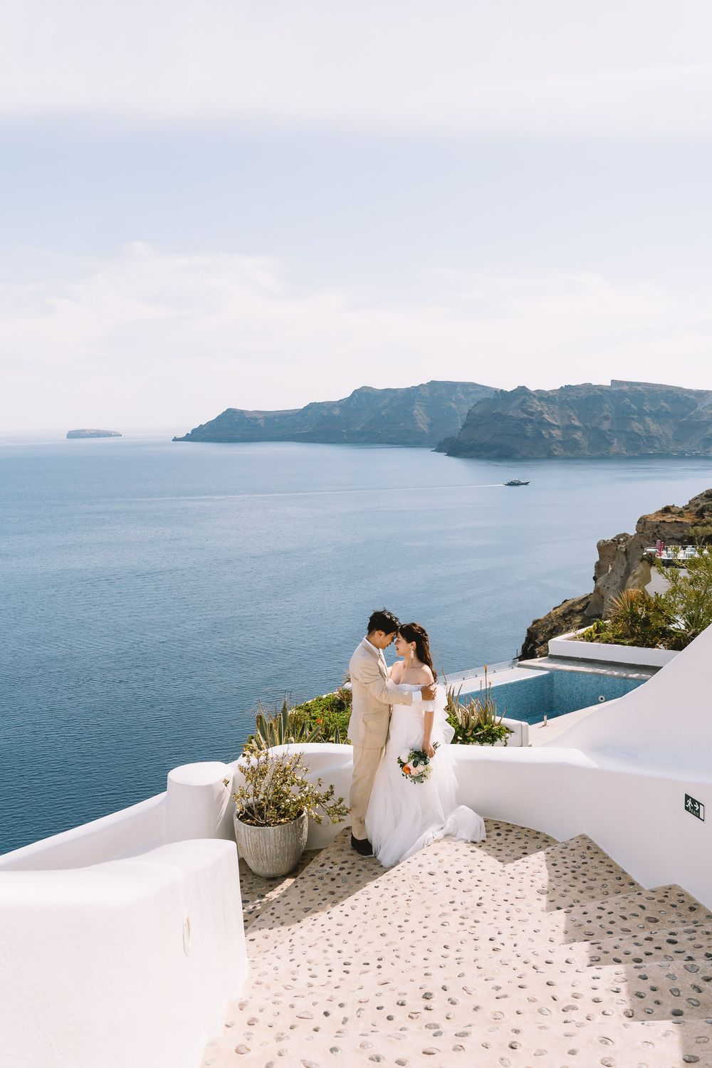 Bride and groom embrace on white steps in Santorini, Greece, with stunning caldera sea views and cliffs behind them.