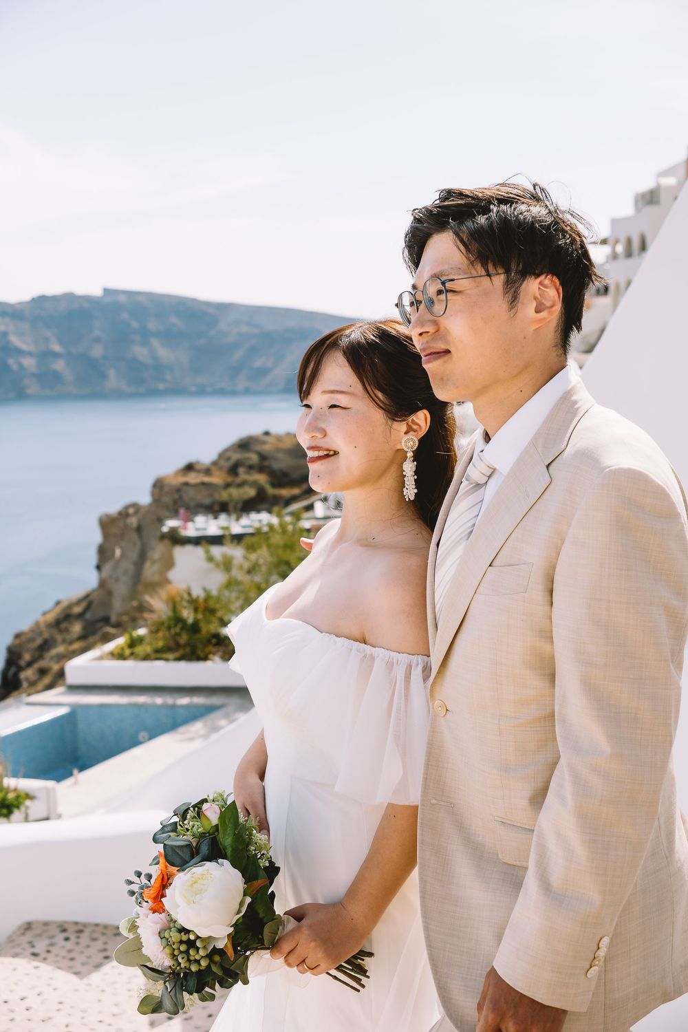 Asian couple in wedding attire posing in Santorini, Greece, with caldera views and white architecture backdrop.