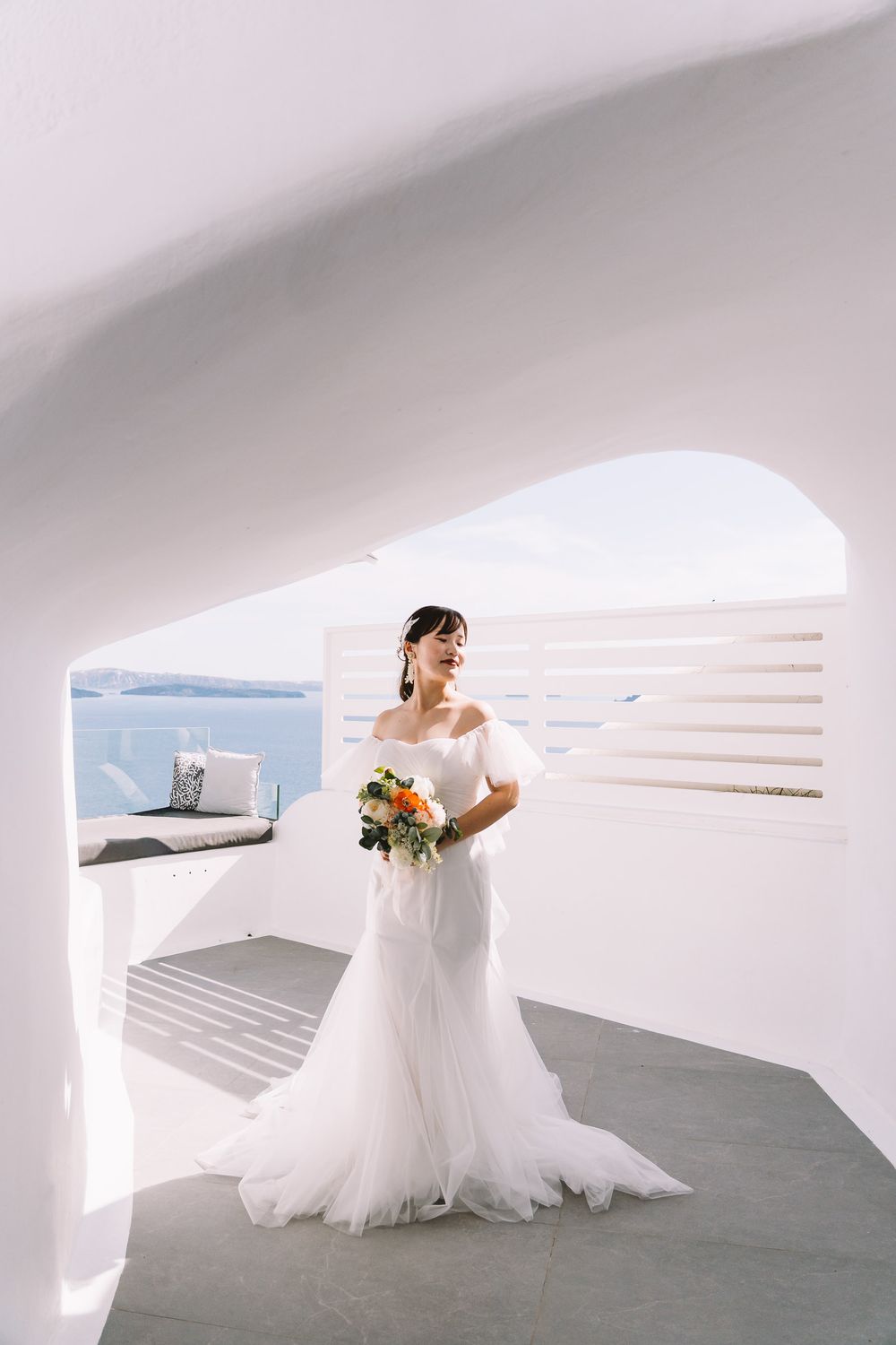 Bride in white off-shoulder gown holds colorful bouquet under arch with Santorini sea view.
