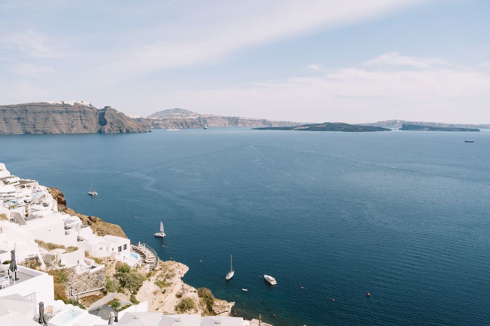 Panoramic view of Santorini caldera with white buildings, sailboats on blue Aegean Sea, and volcanic cliffs, Greece.