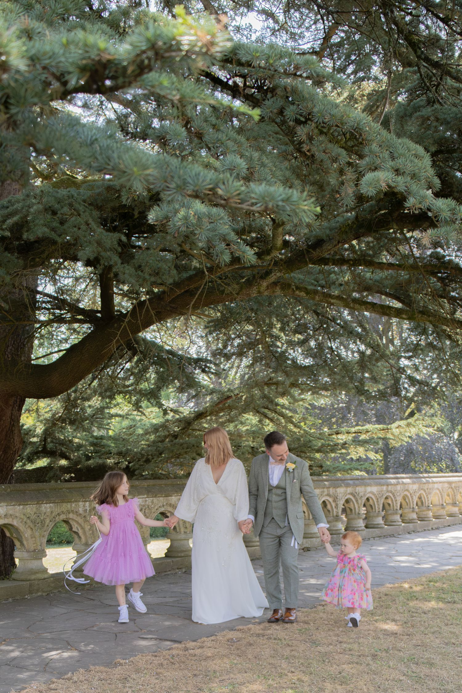 Wedding couple walking hand-in-hand with two young girls in pink dresses under a large cedar tree in a garden.