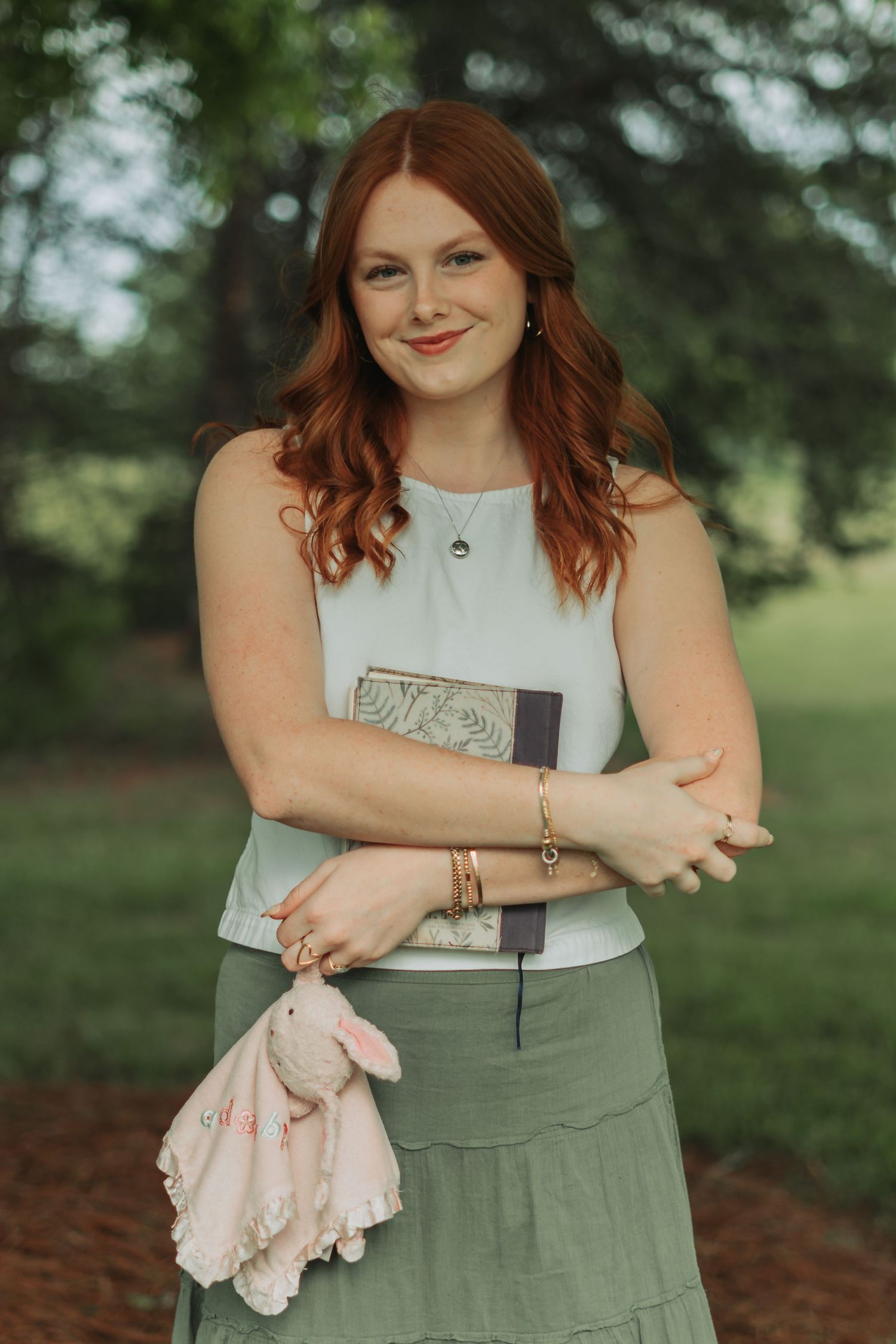 Young redhead woman lying in tall green grass, smiling while reading a book outdoors in a lush meadow.