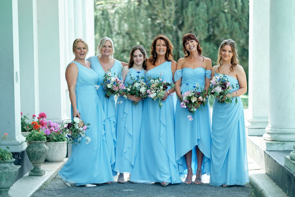 Five bridesmaids in matching light blue gowns holding floral bouquets pose together outdoors near elegant white columns.