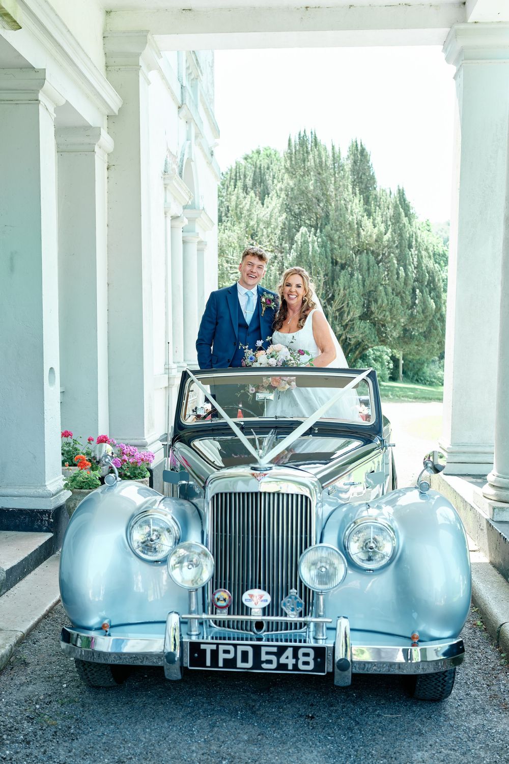 Bride and groom posing behind a vintage silver wedding car with white ribbons outside a grand venue.
