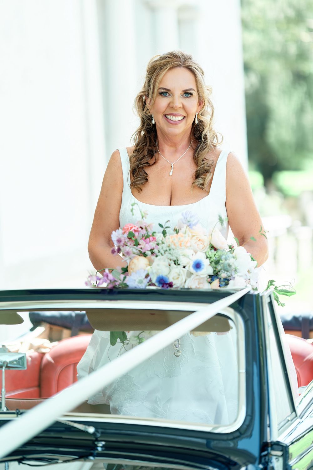 Smiling bride in white gown holding colorful bouquet standing in convertible car on wedding day.