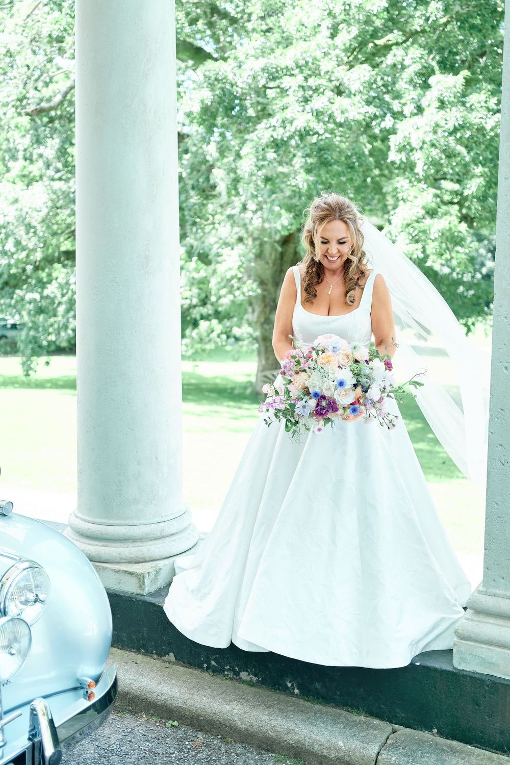 Smiling bride in white ball gown holding colorful bouquet stands between white columns near vintage car outdoors.