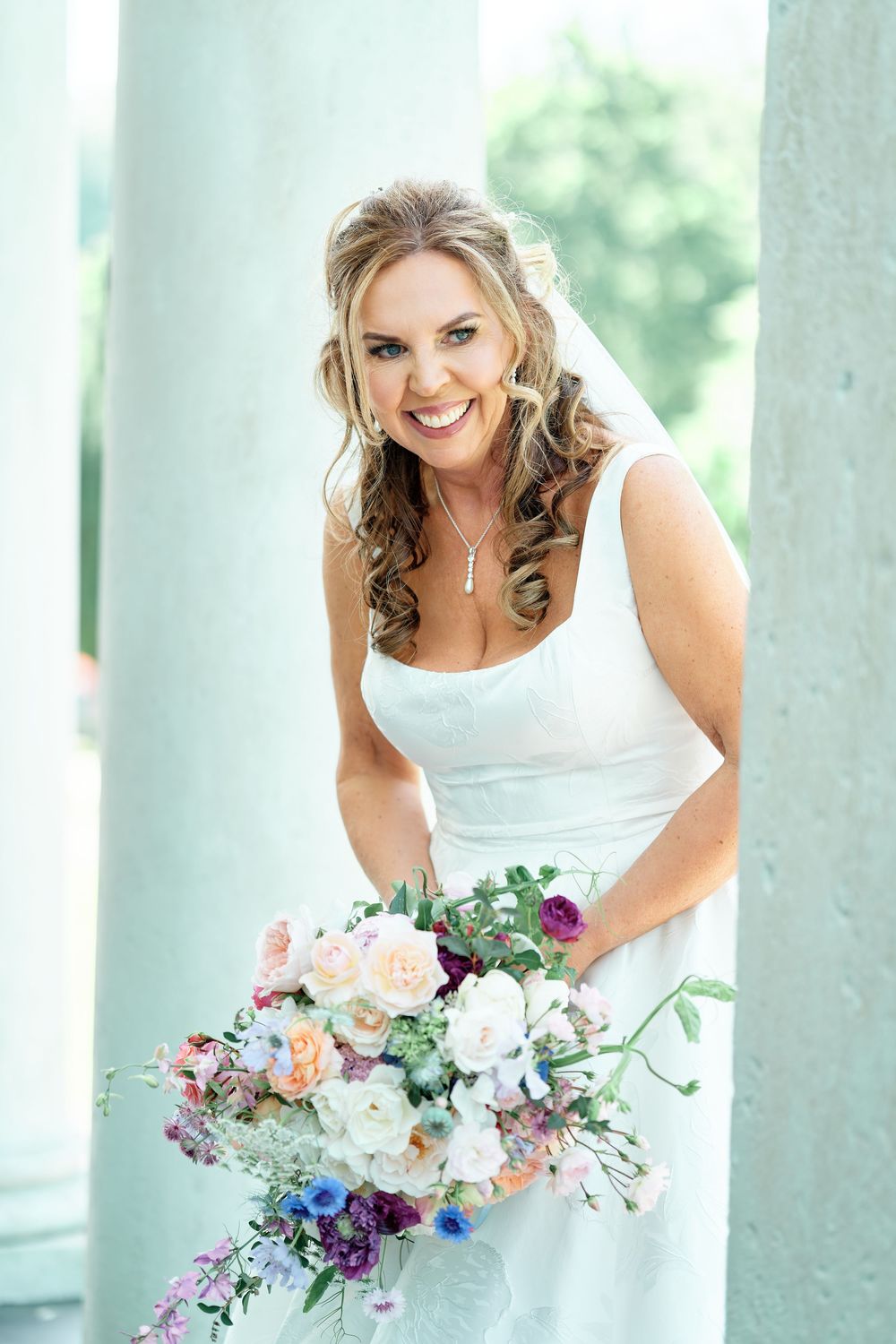 Smiling bride in white dress holding colorful floral bouquet standing between elegant columns on her wedding day.
