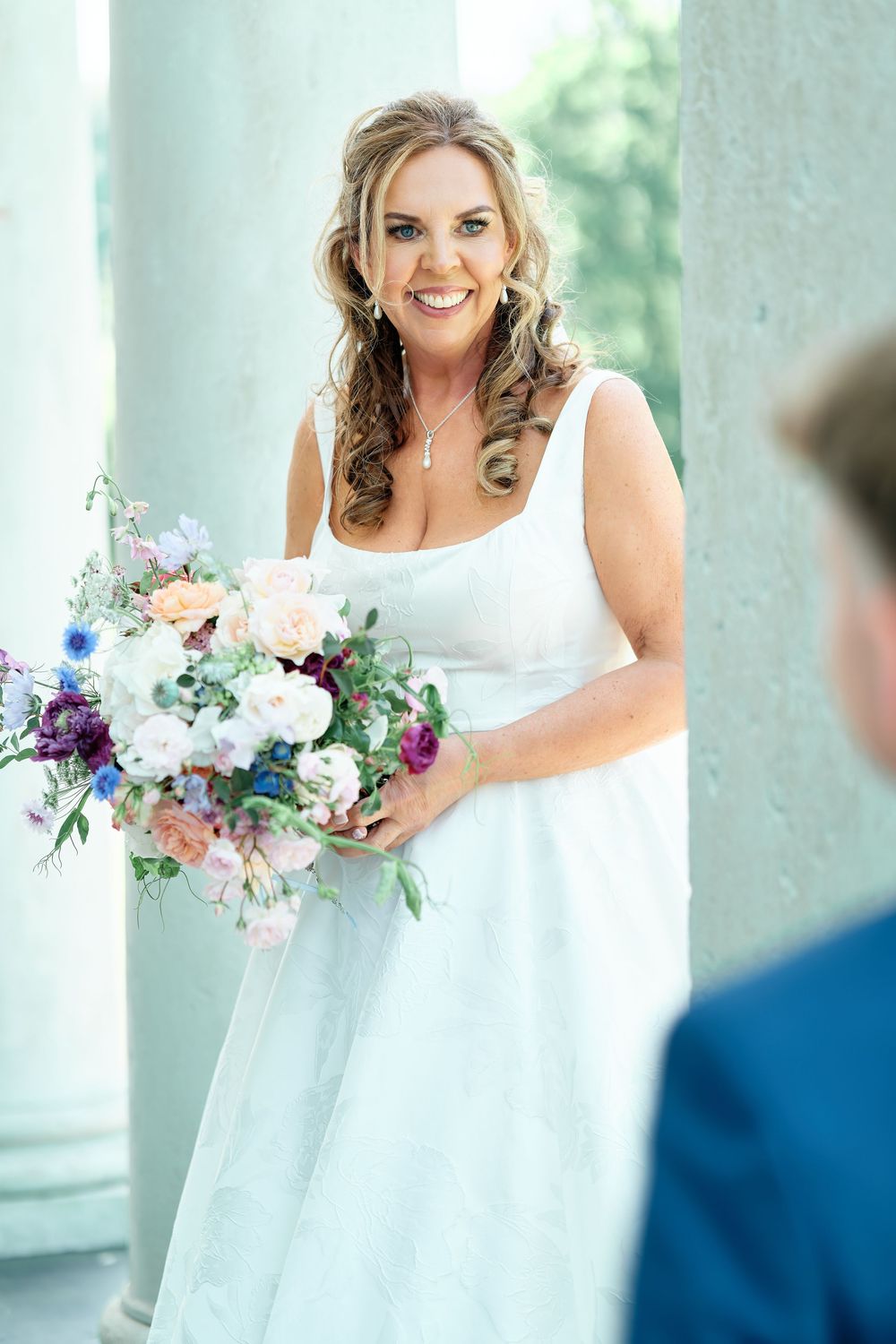 Smiling bride in white gown holds colorful bouquet of purple, pink, and blue flowers beside elegant columns.
