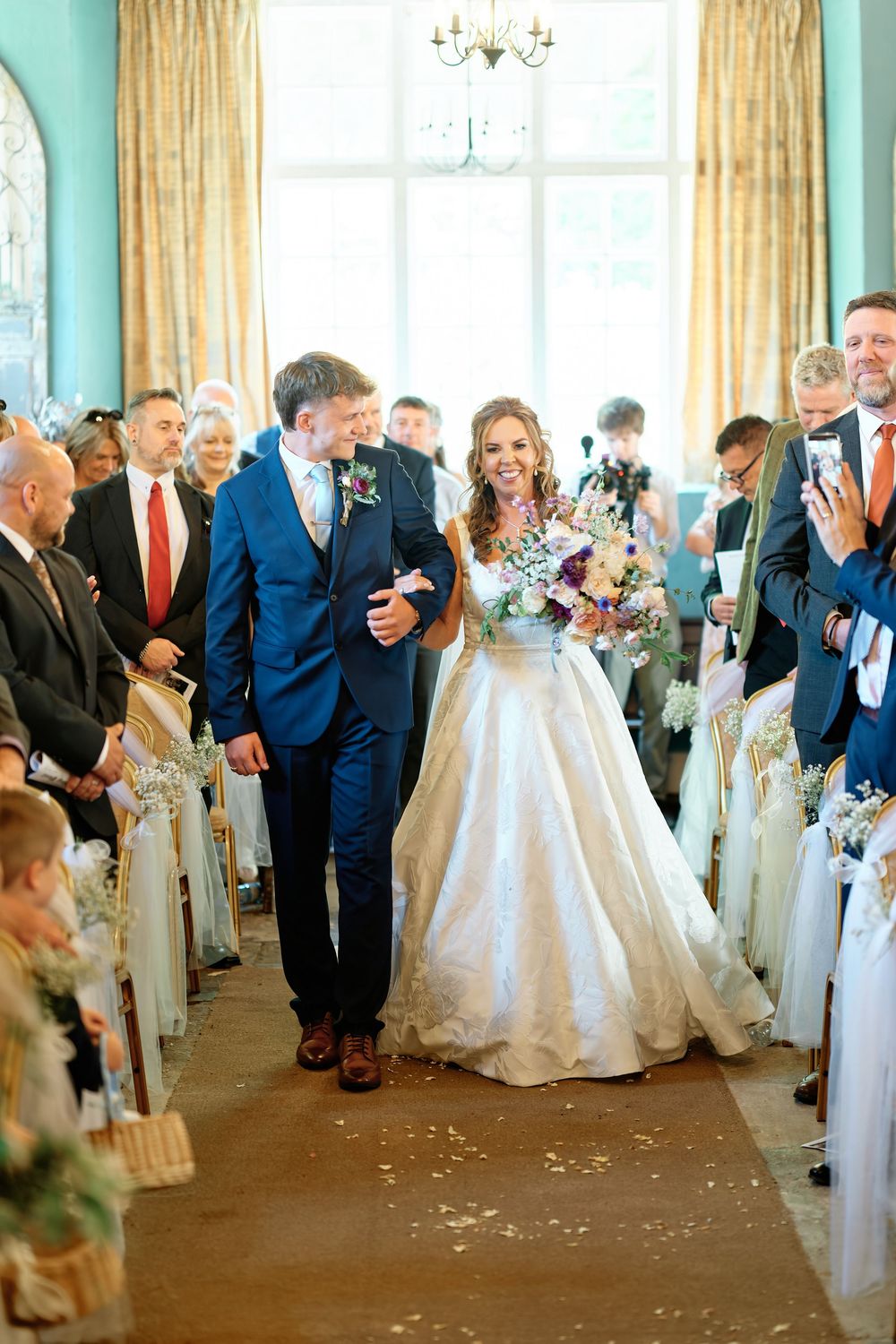 Bride and groom walk down the aisle smiling, guests cheering, elegant blue room with floral bouquet.
