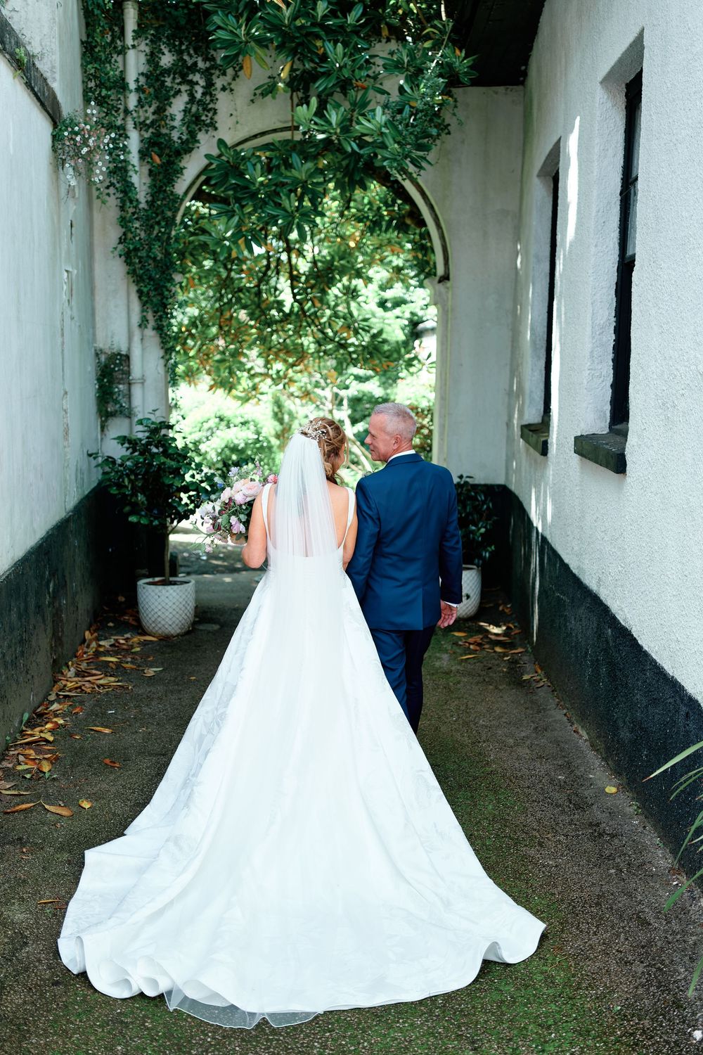 Bride and groom walking hand in hand through a garden archway, bride wearing a long white gown with veil and train.