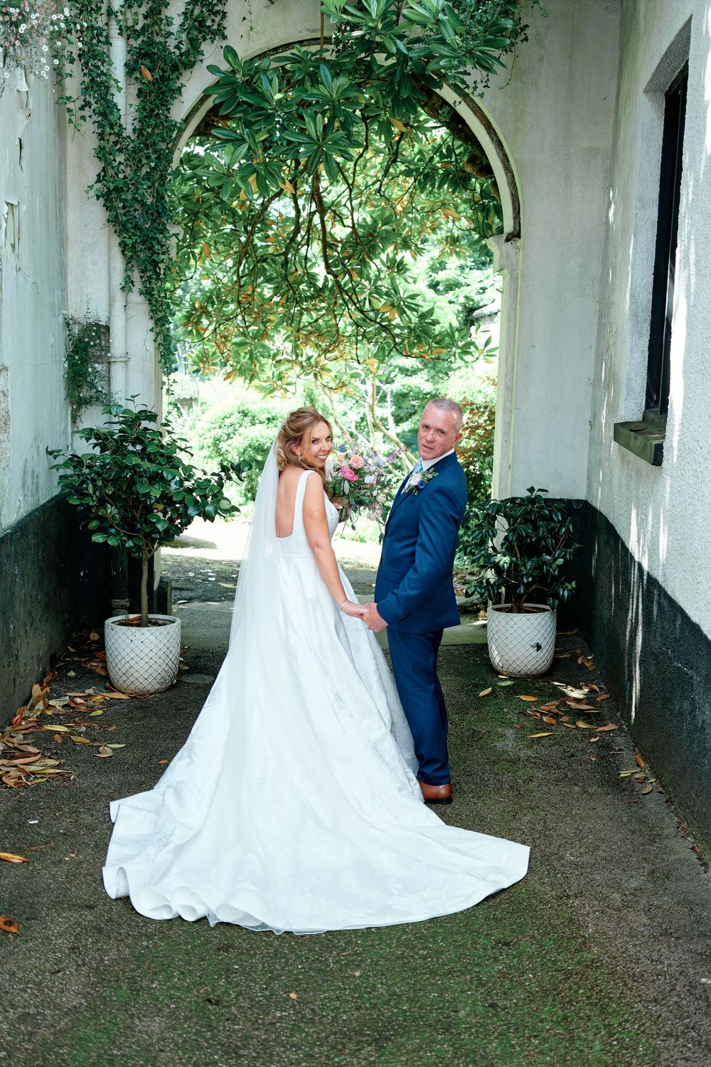 Bride and groom holding hands under a lush garden arch, bride in elegant white gown with long train.