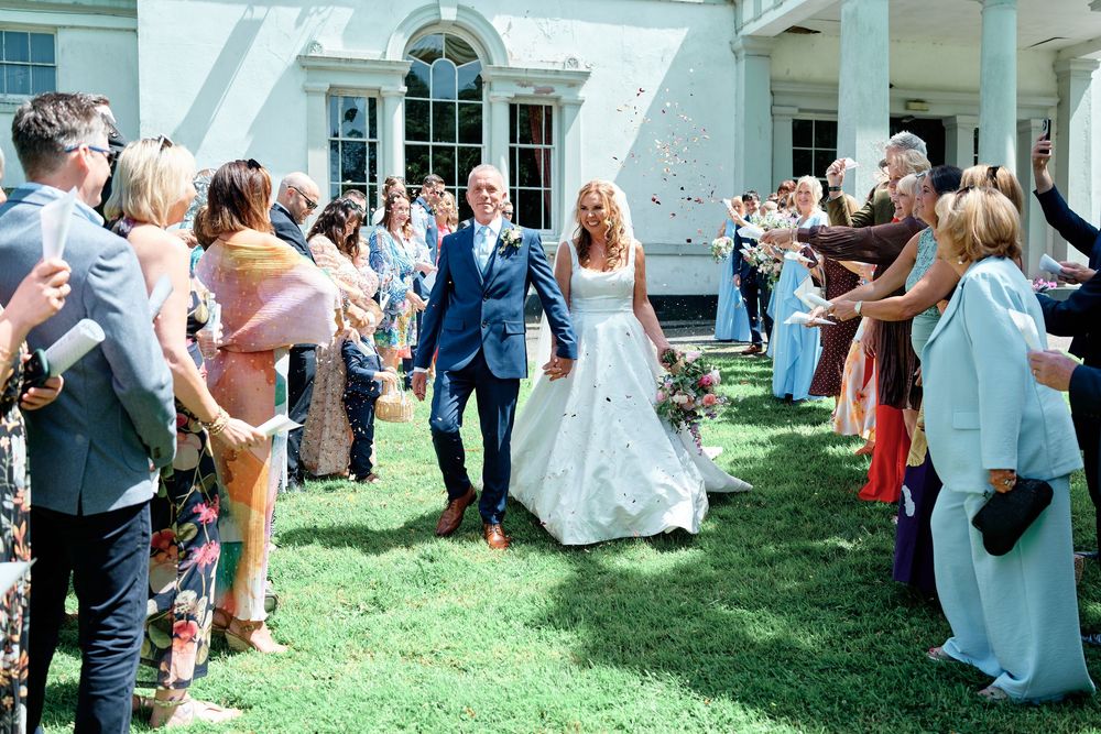 Bride and groom walk through confetti shower surrounded by smiling wedding guests outside elegant white manor house.