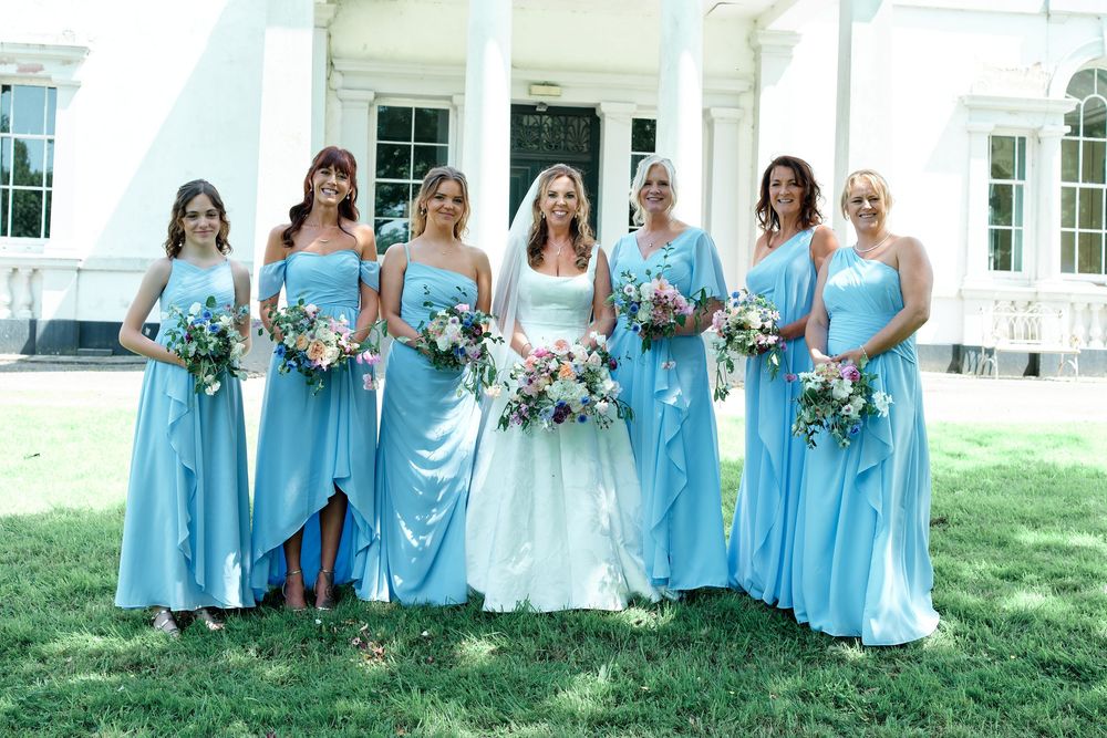 Bride in white gown with six bridesmaids in light blue satin dresses holding bouquets outside elegant white building.