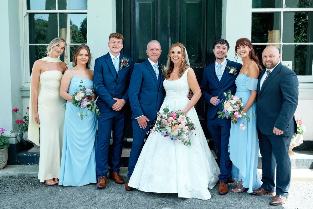 Wedding party posing outdoors, bride in white gown with bouquet, groomsmen in navy suits, bridesmaids in blue dresses.