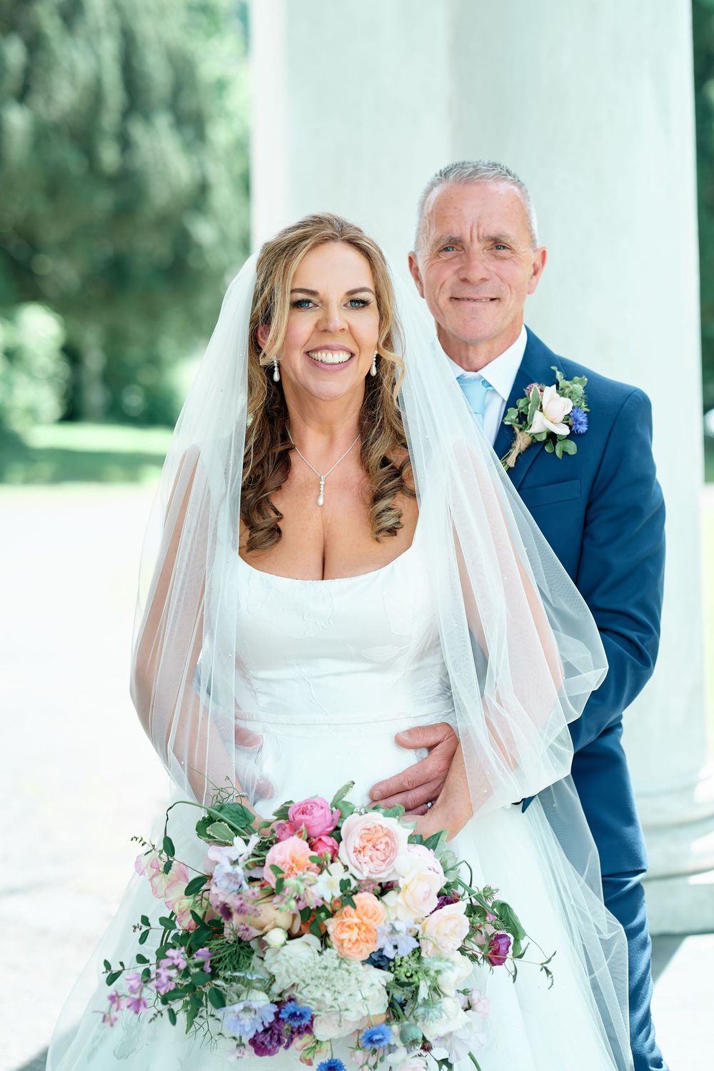 Bride in white gown with veil holding colorful bouquet stands with groom in navy suit at elegant wedding venue.