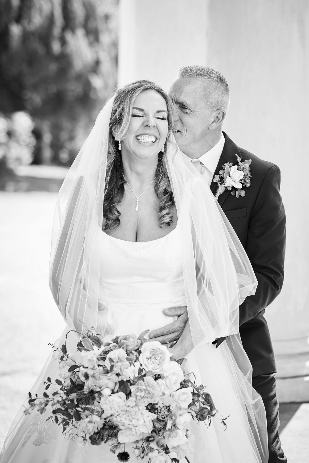 Black and white photo of a laughing bride holding a bouquet while groom kisses her cheek on their wedding day.