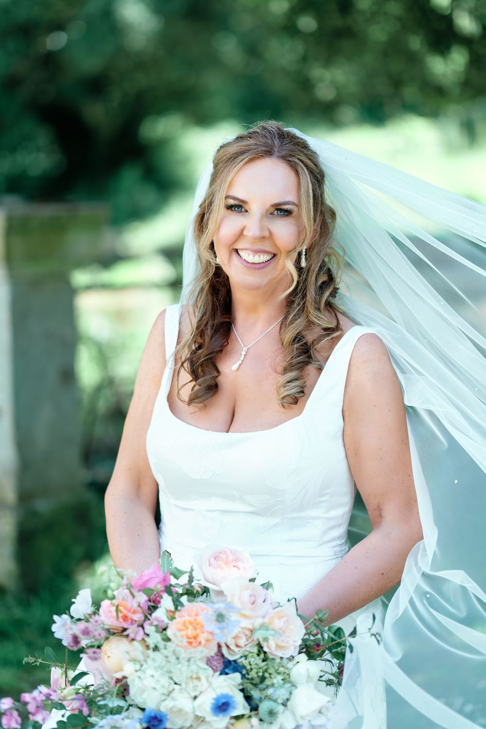 Smiling bride in white gown and veil holds colorful bouquet of pink, blue, and white flowers outdoors.