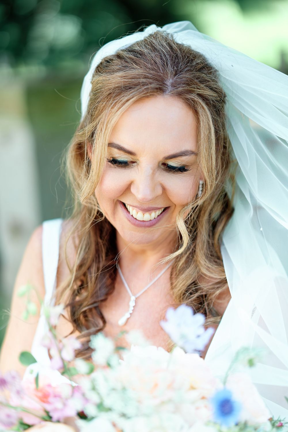 Smiling bride wearing a veil and necklace, holding a colorful bouquet of flowers on her wedding day.