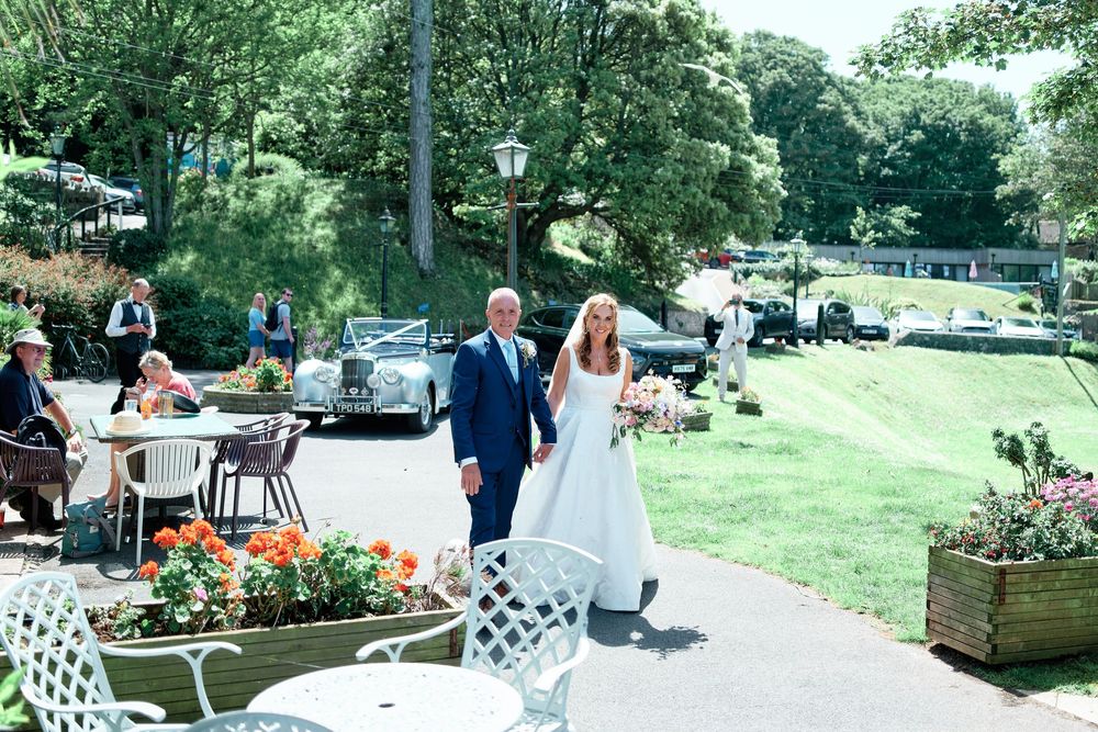 Bride and groom walking through a sunlit garden venue with white furniture and classic cars in the background.