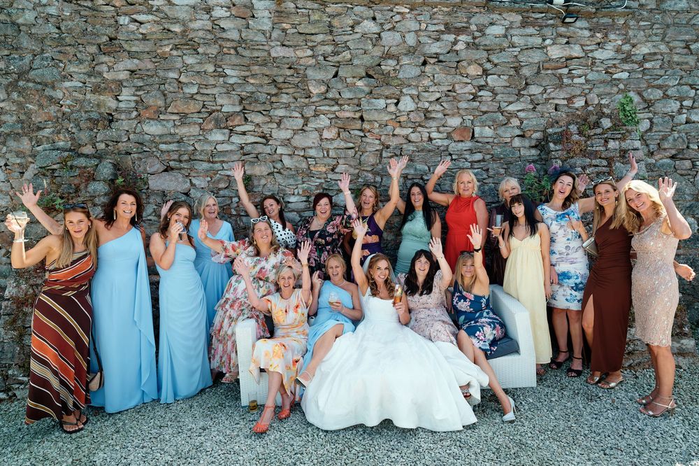Bride in white gown poses with group of women guests in front of stone wall at outdoor wedding celebration.