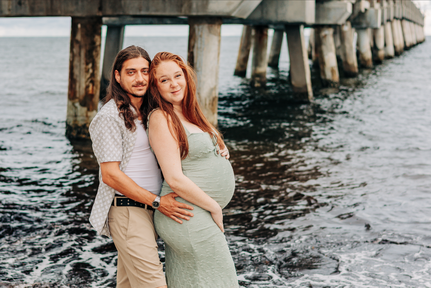 Couple poses by ocean pier, pregnant woman in green dress held by partner, Maternity photoshoot Lake Worth pier