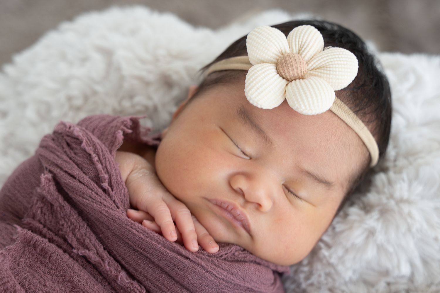 Sleeping newborn wrapped in mauve with a cream flower headband, resting on a soft white blanket.