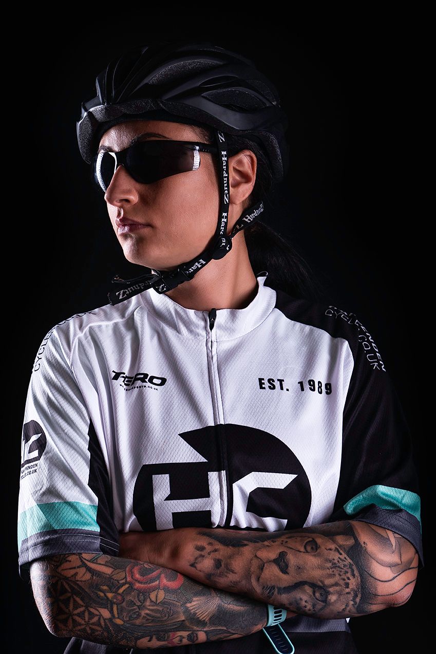 Tattooed female cyclist in black helmet, sunglasses, and white HC team jersey with arms crossed against dark background.