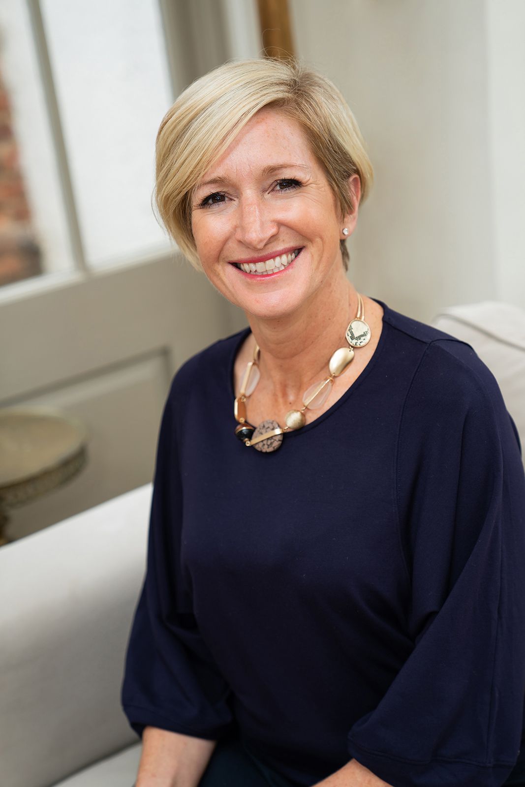 Smiling middle-aged blonde woman in navy blouse with statement necklace seated in bright interior.