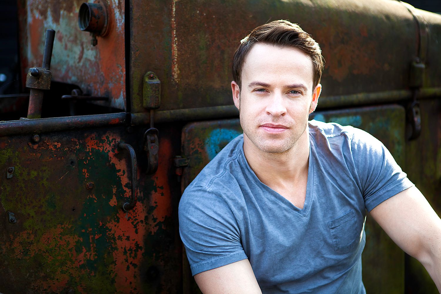 Handsome man in gray t-shirt leaning against rusty metal surface outdoors.