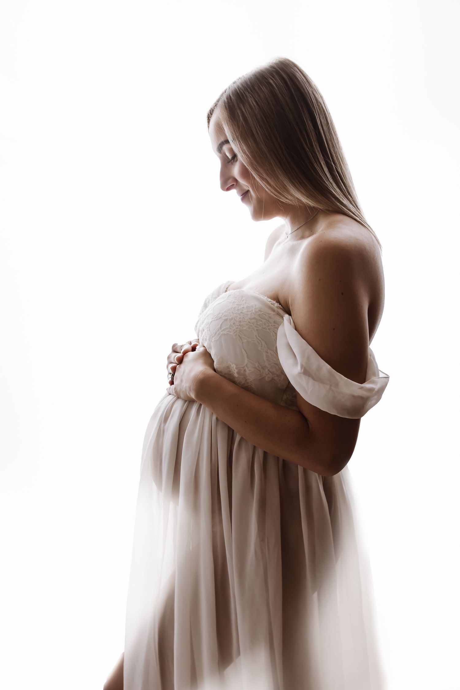 Pregnant woman in elegant beige off-shoulder dress cradling her baby bump, smiling softly against white background.