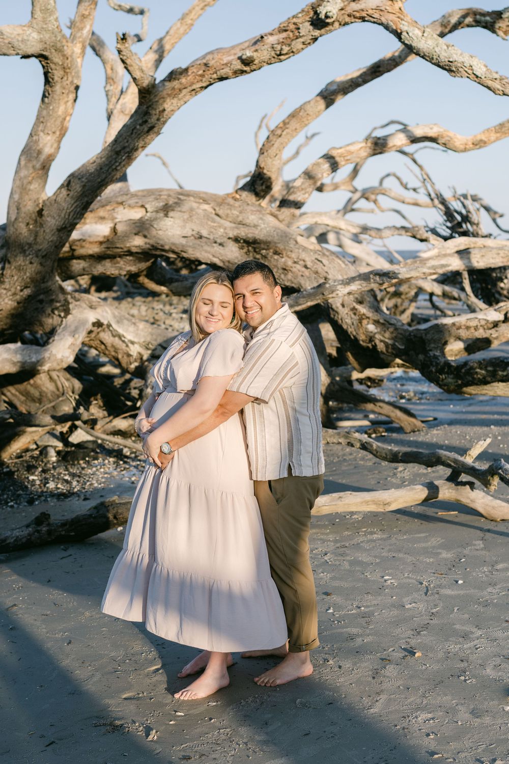 Expectant couple embracing among the driftwood at Jekyll Island’s Driftwood Beach.