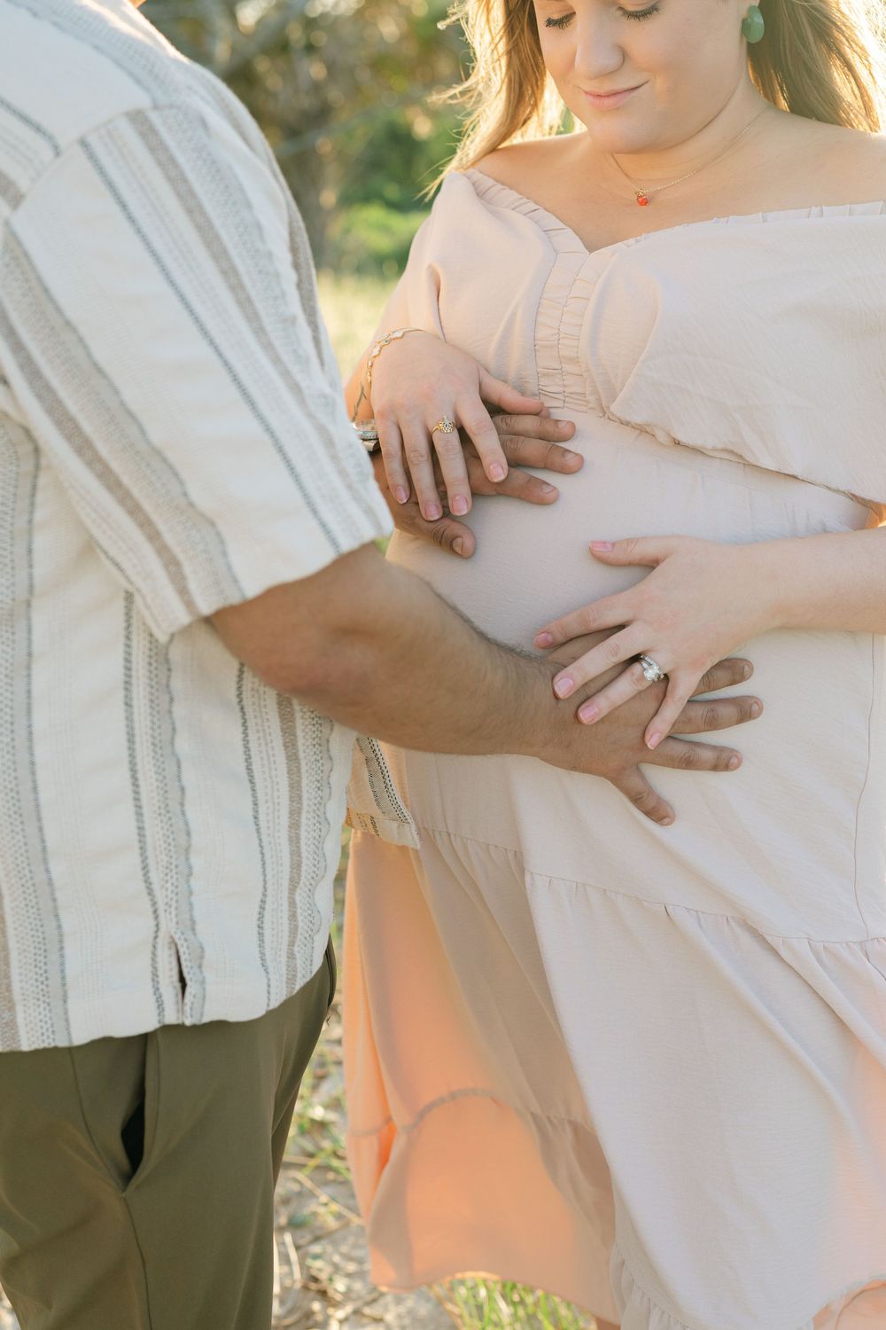 Expectant parents’ hands resting together on her belly in the warm evening light.