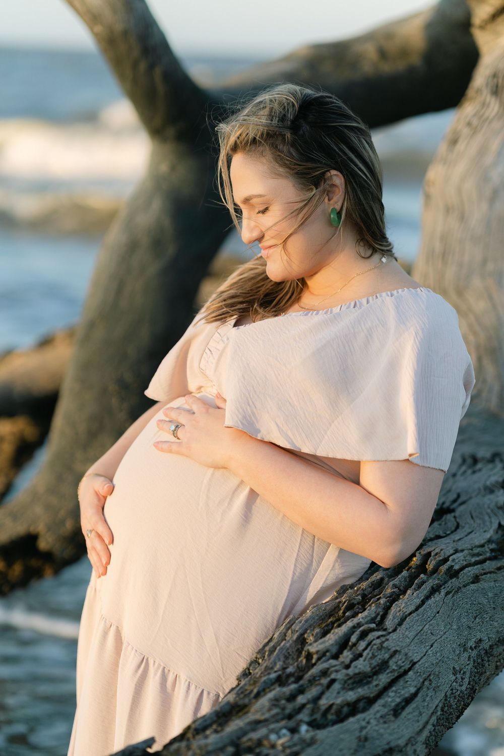 Expectant mother leaning against driftwood and looking down at her belly.