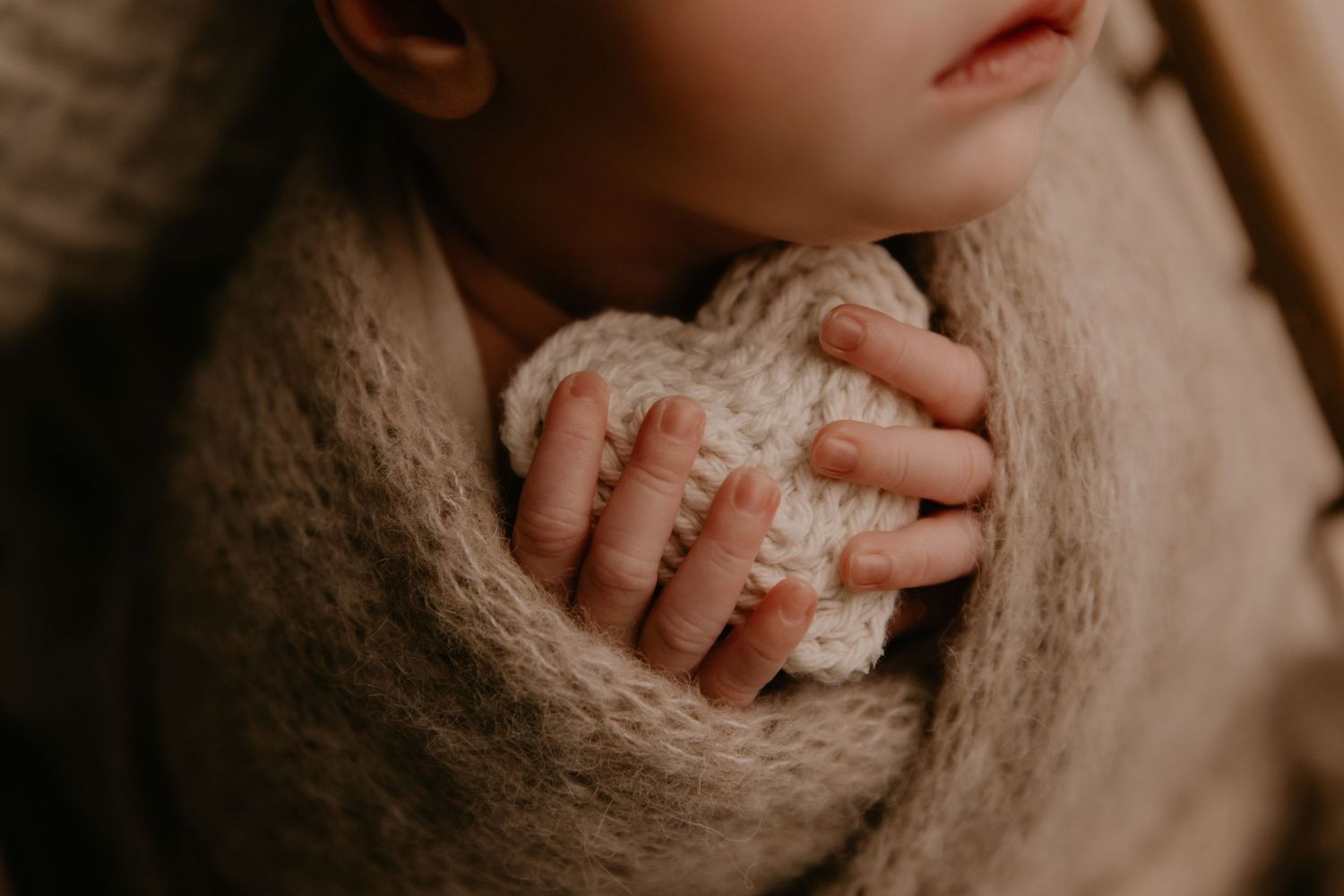 Close-up of a newborn baby wrapped in a soft beige knit blanket, holding a small crocheted heart.