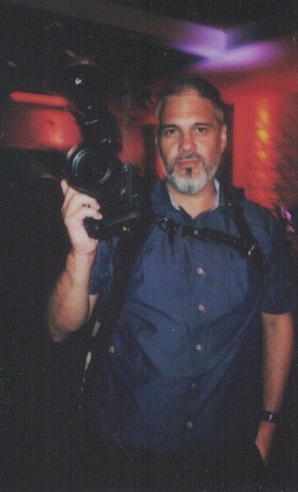 Man holding professional video camera on his shoulder in a dimly lit venue with colorful lighting.