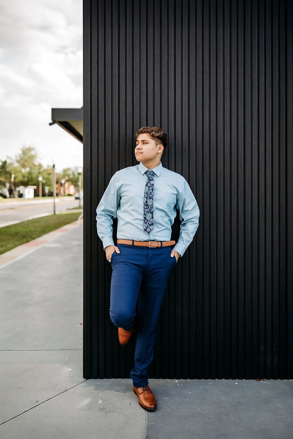 Young man in light blue dress shirt, navy pants, and patterned tie leaning against black wall outdoors.