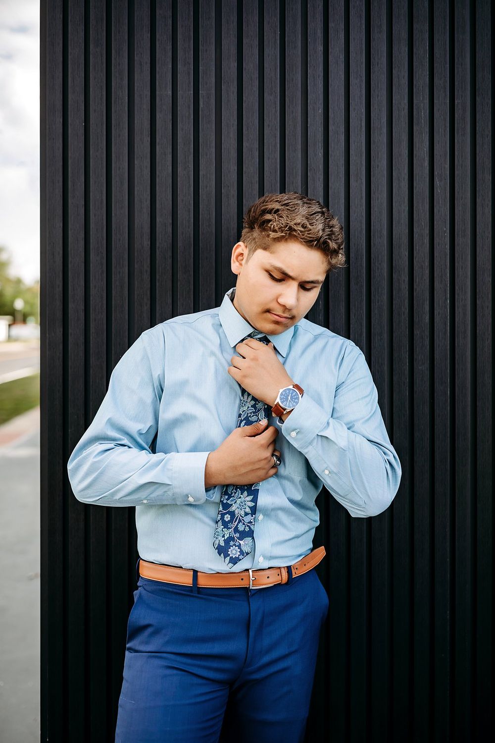 Young man in light blue shirt adjusting floral tie, wearing blue pants and brown belt, standing against black wall.
