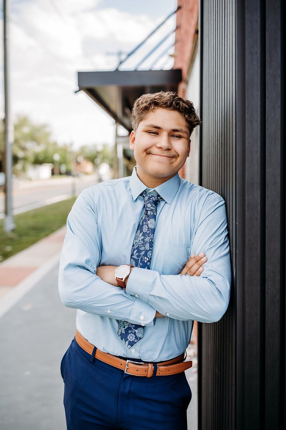 Young man in blue dress shirt and floral tie leans against building with arms crossed, smiling outdoors.