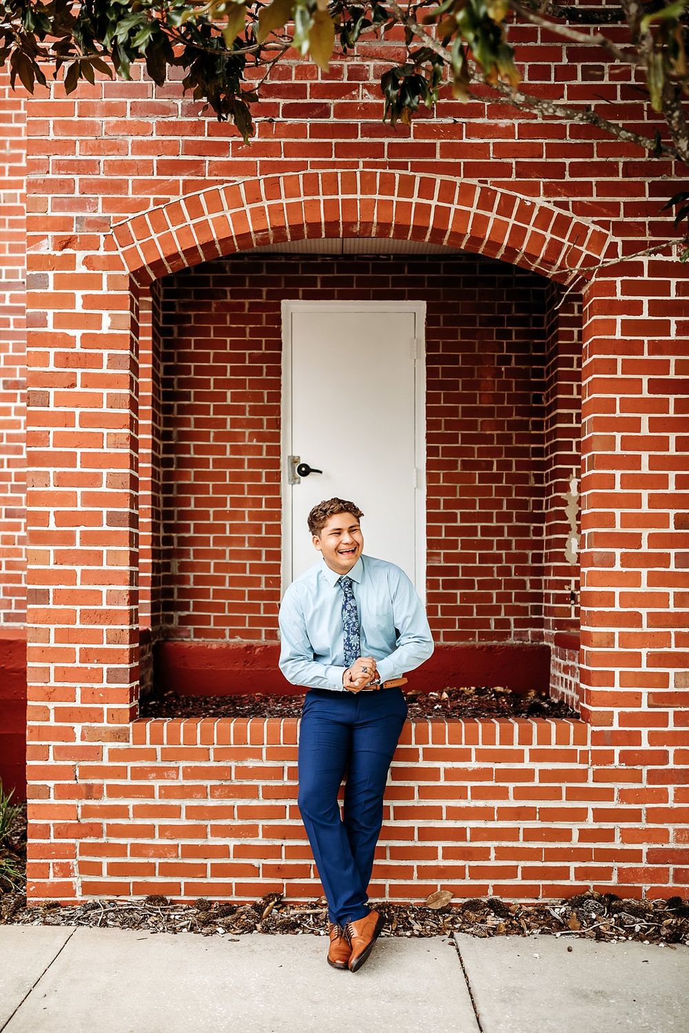 Young man in blue suit and floral tie smiling against a red brick wall with arched doorway.