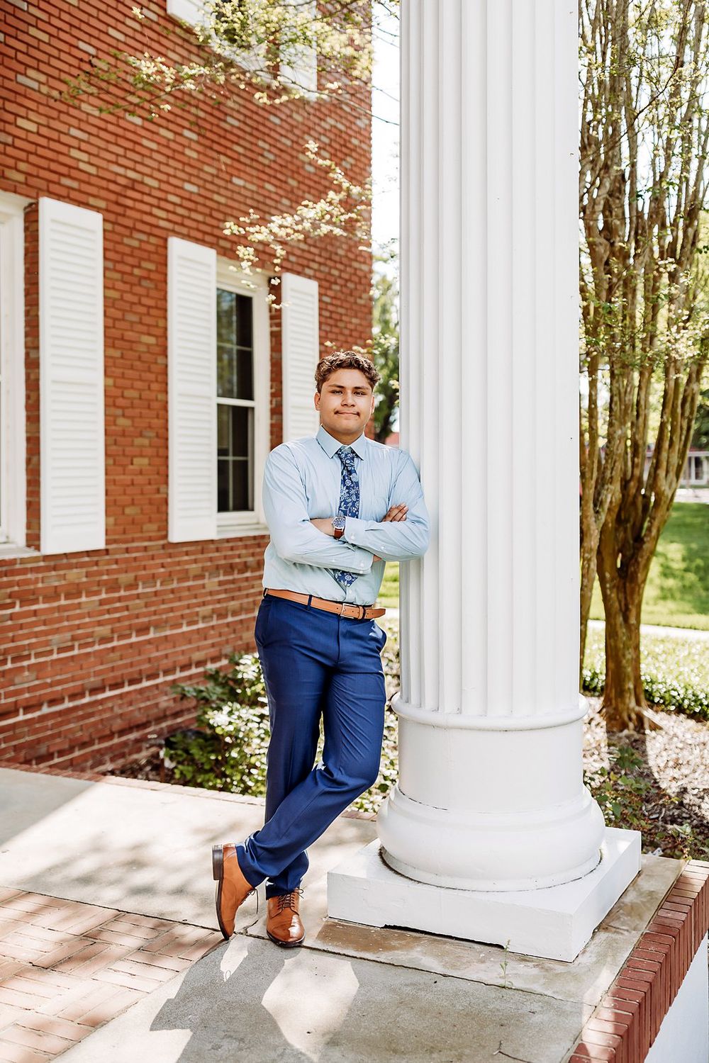 Young man in blue suit and tie leaning against white column outside brick building, arms crossed, smiling.