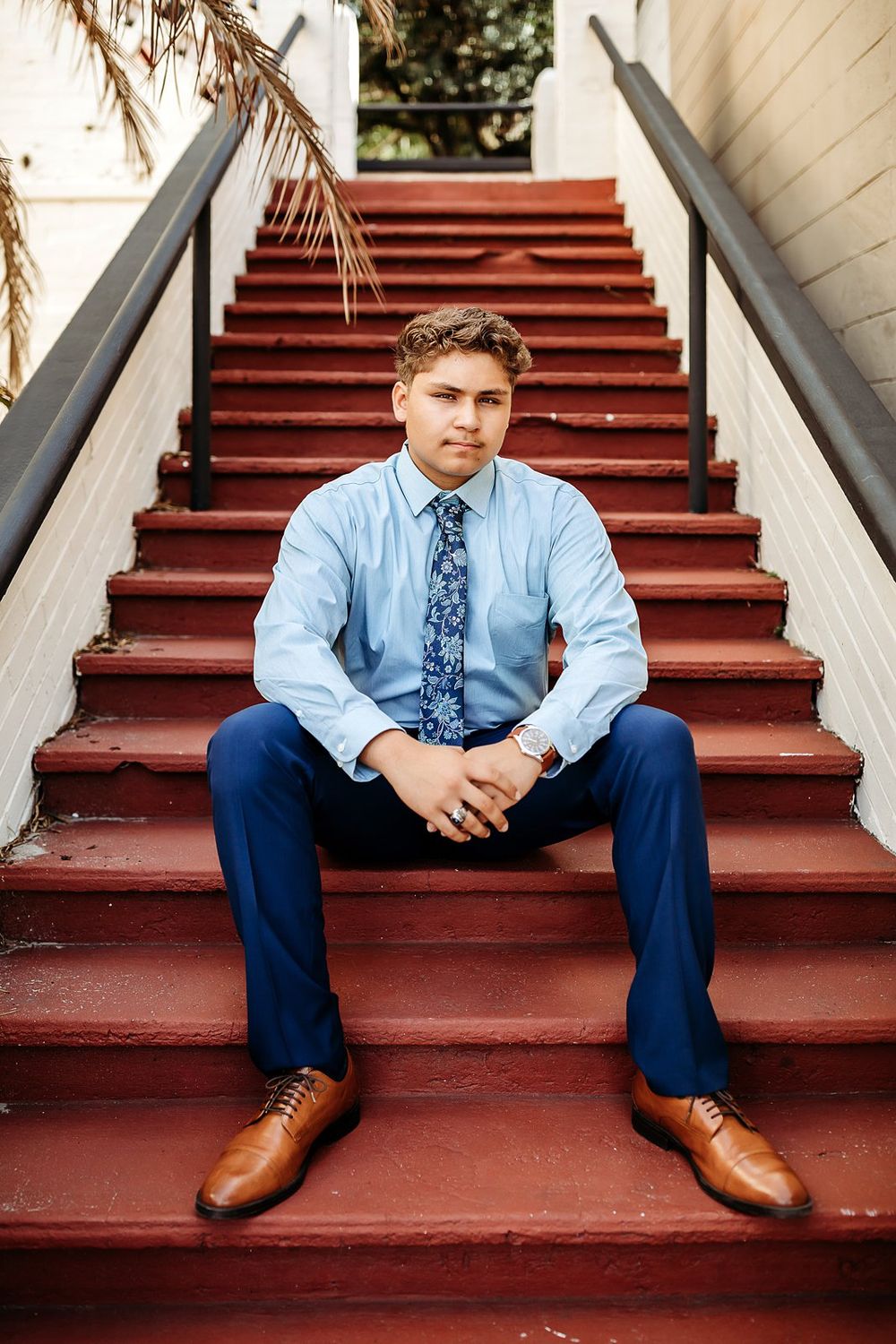 Young man in blue shirt, floral tie, and navy pants sitting on red stairs wearing brown oxford shoes.