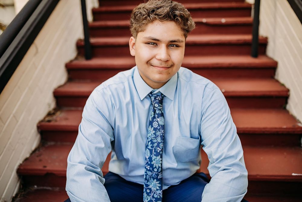 Young man in blue shirt and floral tie smiling on red brick stairs in a professional portrait photo.