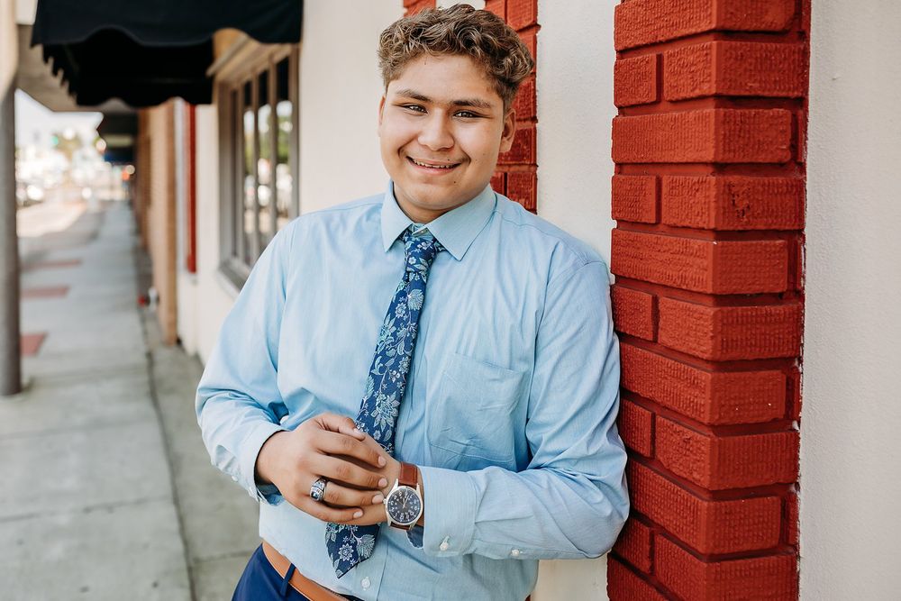 Young man in light blue dress shirt and floral tie leaning against red brick wall on a sidewalk.