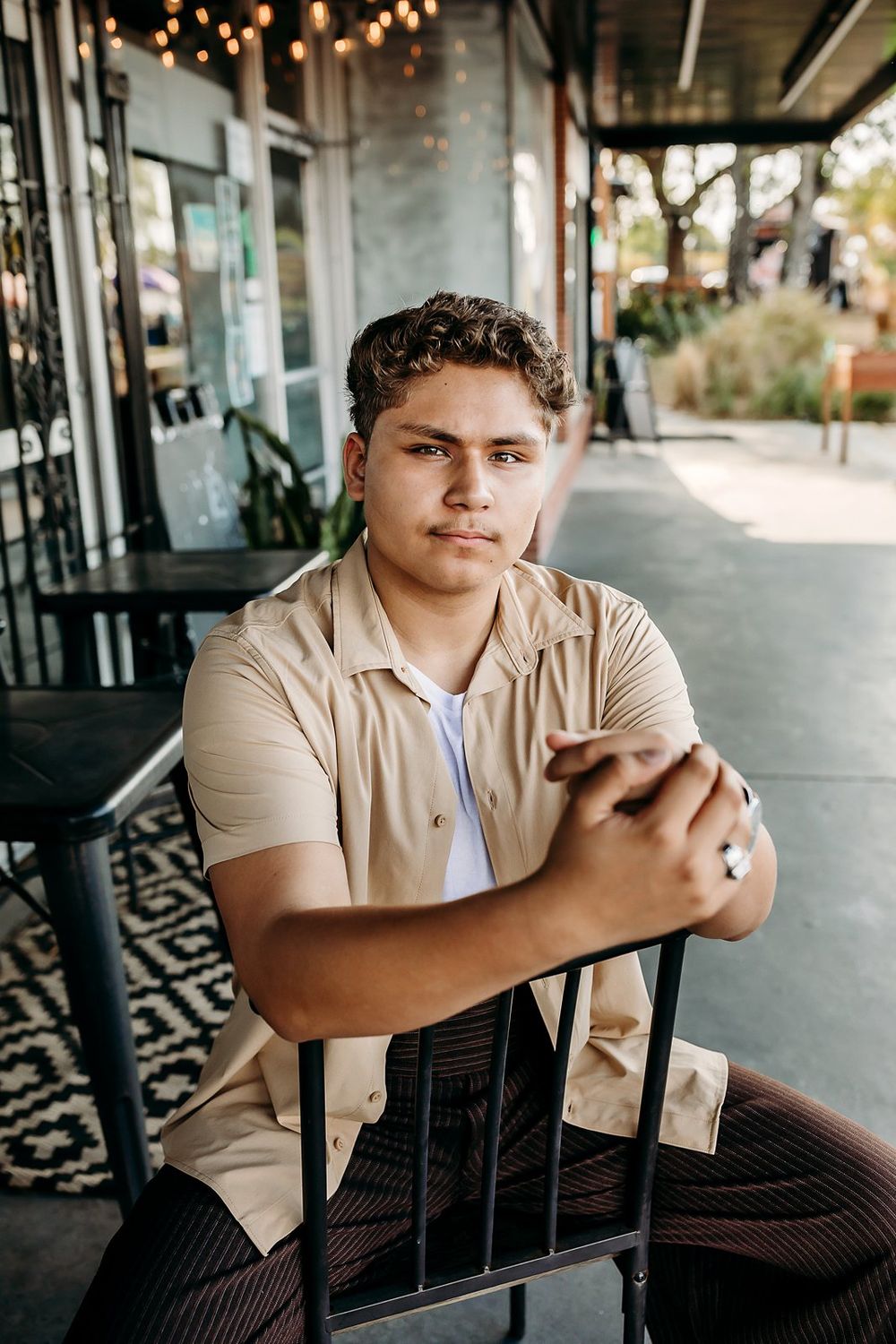 Young man in beige shirt sits confidently at outdoor cafe table, arms crossed on chair back.