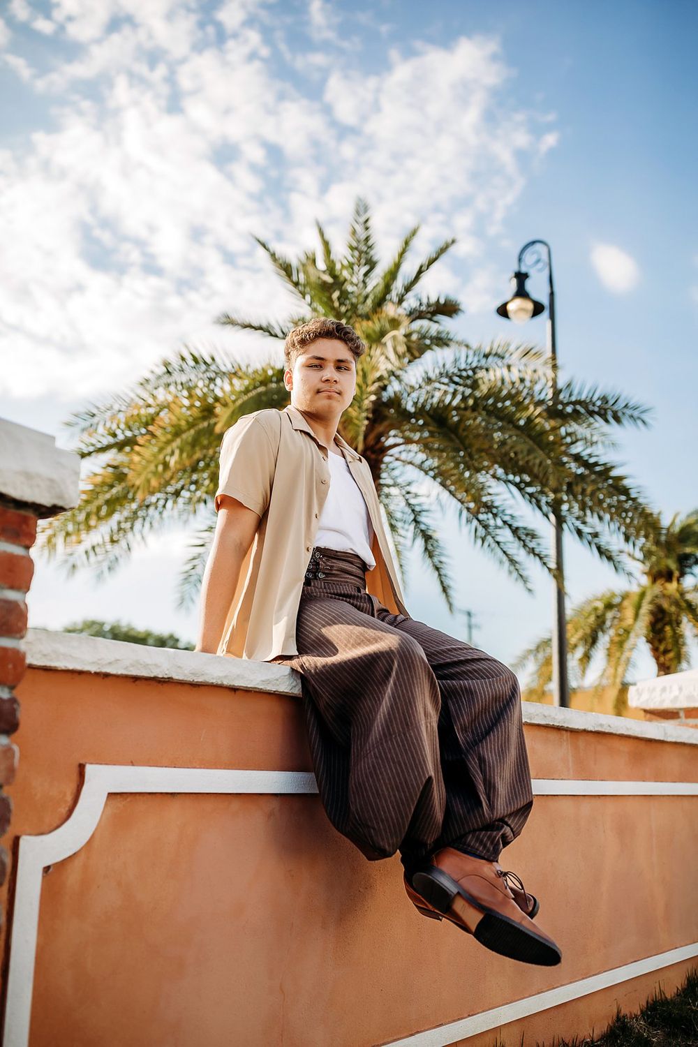 Young man in beige shirt and striped trousers sitting on a wall with palm trees and blue sky.